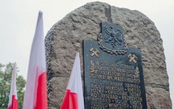Kamienny obelisk na bliskim kadrze. W tle flagi Polski.