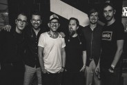 czarno-białe zdjęcie przedstawiające sześciu członków zespołu Happysad, stojących przodem do…
