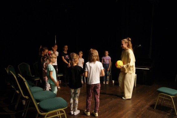 Na scenie, prowadząca ubrana w jasny strój, trzymając w rękach piłkę zwraca się dogrupki dzieci.