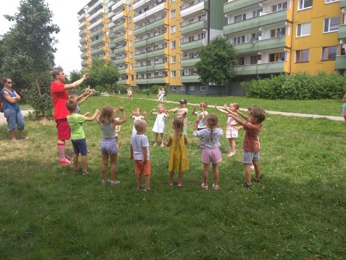 Stojące na trawie w kółku dzieci, naśladujące ruchy stojącego z nimi aktora. Wszyscy mają…