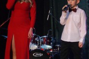 Daria Wantuła w długiej, czerwonej sukni oraz Szymon Wantuła z mikrofonami w rękach na scenie…