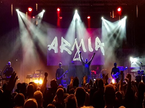 Koncert zespołu Armia. Na pierwszym planie fani, na drugim muzycy na scenie, w tle światła…
