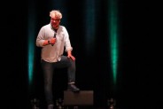 Satyryk Piotr Bałtroczyk z mikrofonem z ręce stoi na scenie, z nogą na odsłuchu. Ma jasną koszulę…