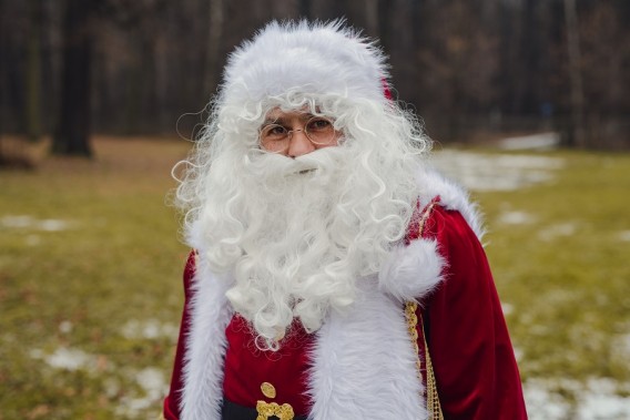 Na tle drzew i trawy stojący Mikołaj z białą brodą, w okularach,w czerwonym stroju ze złotymi i…
