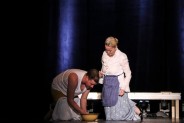 Aktor grający Hanika, ubrany w biały podkoszulek, klęczy przed aktorką grającą jego matkę i myje…