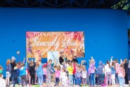 Na tle niebieskiej ściany amfiteatru widok na grupę dzieci biorących udziałw spektaklu. Za nimi…