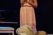 Wokalistka stoi na scenie w różowej sukience i śpiewa do mirofonu. Przed nią leżą zabawki, misie…