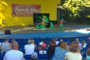 Na scenie amfiteatu  skrzaty Frędzelka i Wiercipietka  w kolorowych kostiumach na tle scenografii…