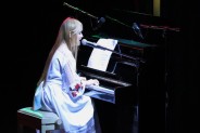 Taisiia Popova w sługiej białej sukni siedzi przy pianinie i śpiewa. Ma długie, jasne włosy
