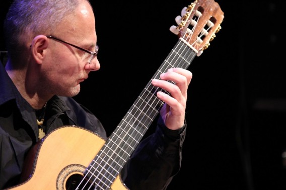 Mężczyzna w średnim wieku w okularach, ubrany w czarną koszulę grający na gitarze klasycznej.