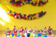 Na żółtym tle sceny amfiteatru wiszą kolorowe balony. Na scenie występuje dziecięcy zespół taneczny