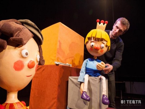 Na pierwszym planie dwie ogromne lalki teatralne. jedna siedzi na szarym klocku, animowana przez…