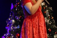Wokalistka w czerwonej sukience stoi na scenie trzymając w ręce mikrofon. Za nią choinka.