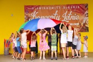 Dzieci bawią się w grupie, na scenie amfiteatru podczas niedzielnych animacji. W tle żółta ściana…