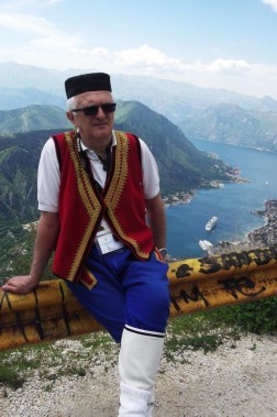 Na pierwszym planie mężczyzna w bałkańskim stroju ludowym. W tle zatoka i góry