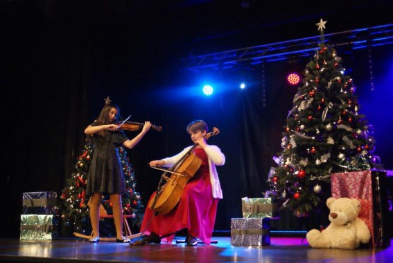 Dwie dziewczyny na scenie. Jedna gra na skrzypcach, druga na wiolonczeli. Obok choinki i prezenty.