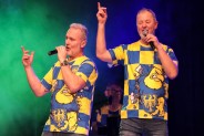 Dwóch mężczyzn śpiewa do mikrofonów na scenie. Oboje mają niebiesko- żółte koszulki