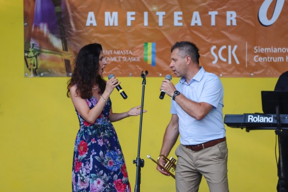 Aleksandra Turek i Jacek Krupa z mikrofonami w dłoniach śpiewają na scenie amfiteatru. W tle na…