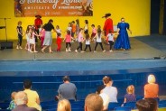 Na niebieskiej scenie, na tle żółtej ściany amfiteatru bawią się dzieci oraz ubrani kolorowe…
