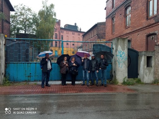 Grafika przedstawia grupę ludzi, stojących w deszczu z parasolami pod bramą dawnego browaru w…