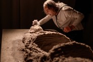 Aktor w białej koszuli pochylający się nad figurą z piasku
