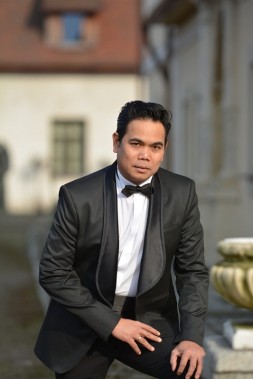 Na zdjęciu wokalista Alexander Martinez ubrany w czarny smoking, muszkę i białą koszulę na tle…