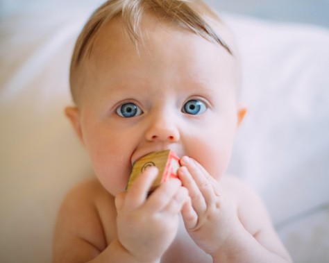 Zbliżenie na buzię małego dziecka z klockiem w ustach. Maluch ma jasne włosy i niebieskie oczy