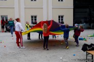Dzieci bawiące się na dziedzincu pałacu, podrzucają kolorowe piłeczki przy pomocy kolorowej płachty