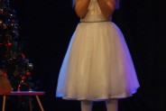 Wokalistka w białej sukience stoi na scenie trzymając w ręce mikrofon. Za nią choinka i konik na…