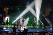 Na scenie Amfiteatru trzej muzycy grający na gitarach. Kolorowe światła z widocznymi białymi…