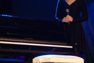 Wokalistka ubrana w czarną sukienkę stoi za pianinem, trzymając z ręce mikrofon