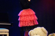 Wokalistka stoi na scenie w czarnej bluzce i czerwonej spódniczce. Śpiewa do trzymanego w ręku…