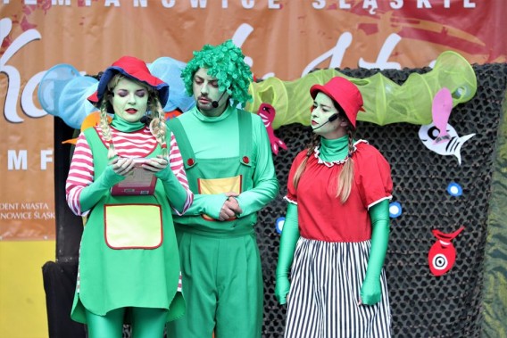Aktorzy przebrani w trzy zielone utopce stoją na tle scenografii na scenie amfiteatru