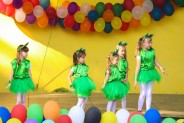 Na żółtym tle sceny wiszą kolorowe balony. Na scenie występuje dziecięcy zespół taneczny. dzieci…